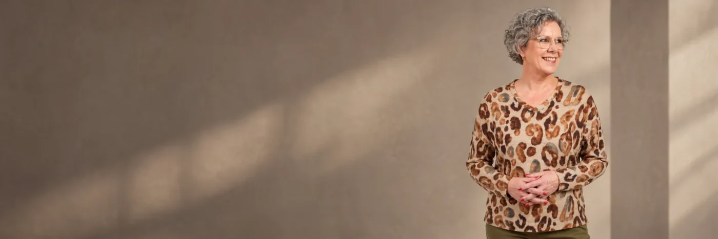Lachende oudere vrouw met grijs krullend haar in een beige top met dierenprint, voor een blog over goede kledingmerken voor oudere dames.